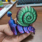 Colorful articulated 3D printed snail toy with a green shell and purple body, held in a hand